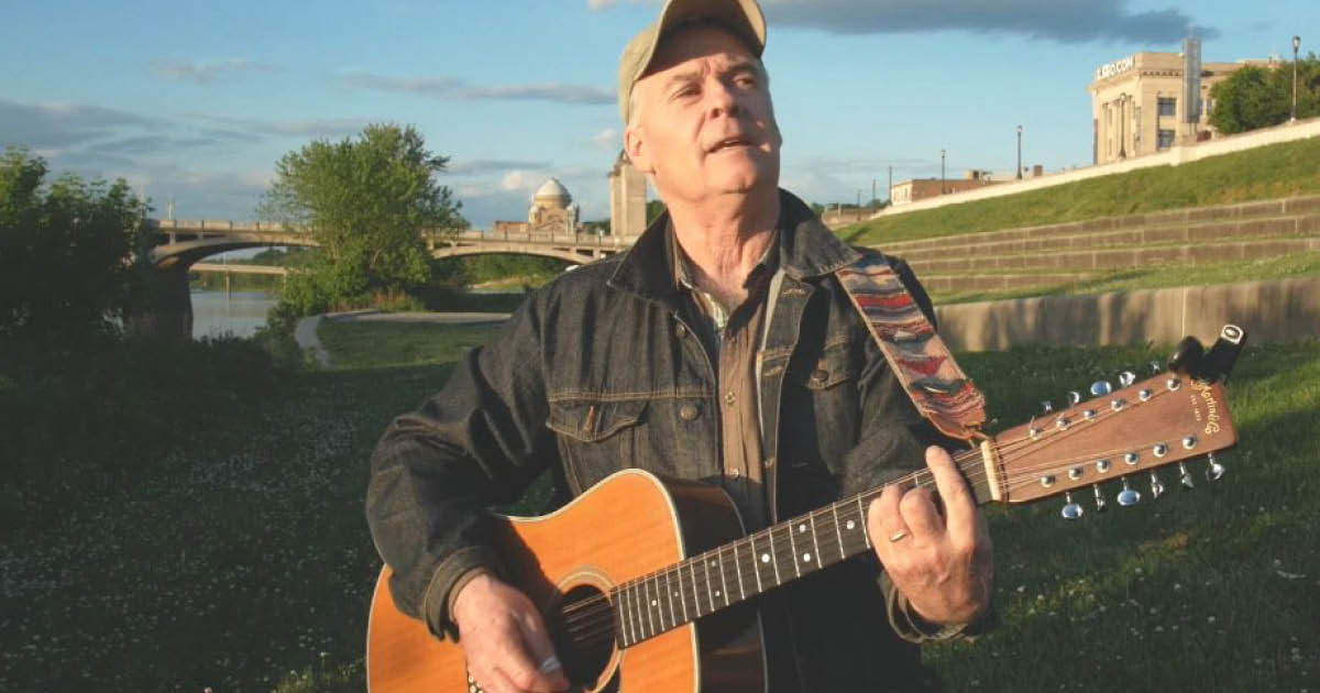 Folksinger Don Shappelle plays his guitar on a lawn next to a river