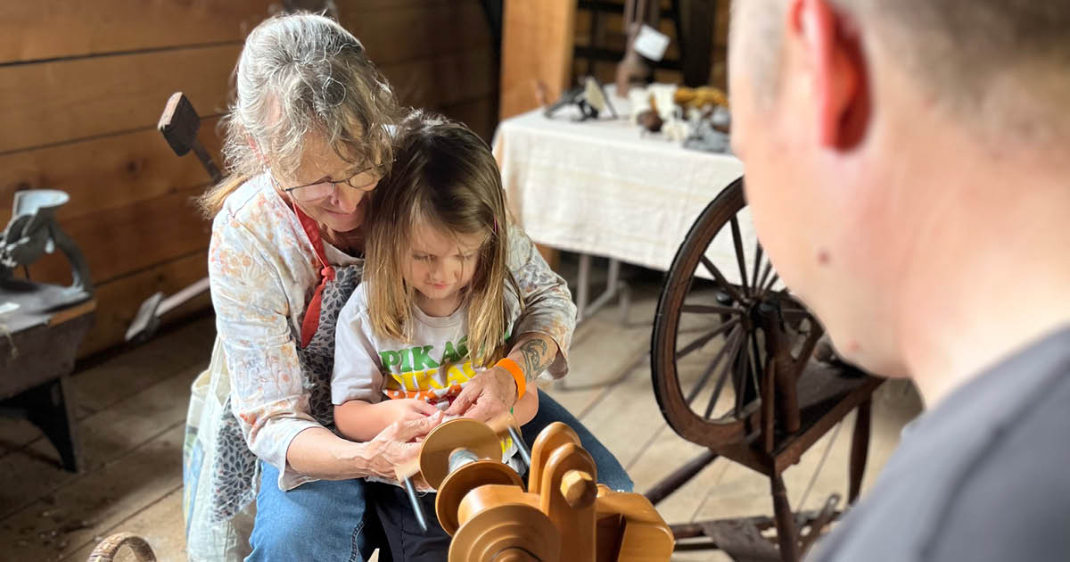 A weaver demonstrating for a young girl on her lap how to spin yarn